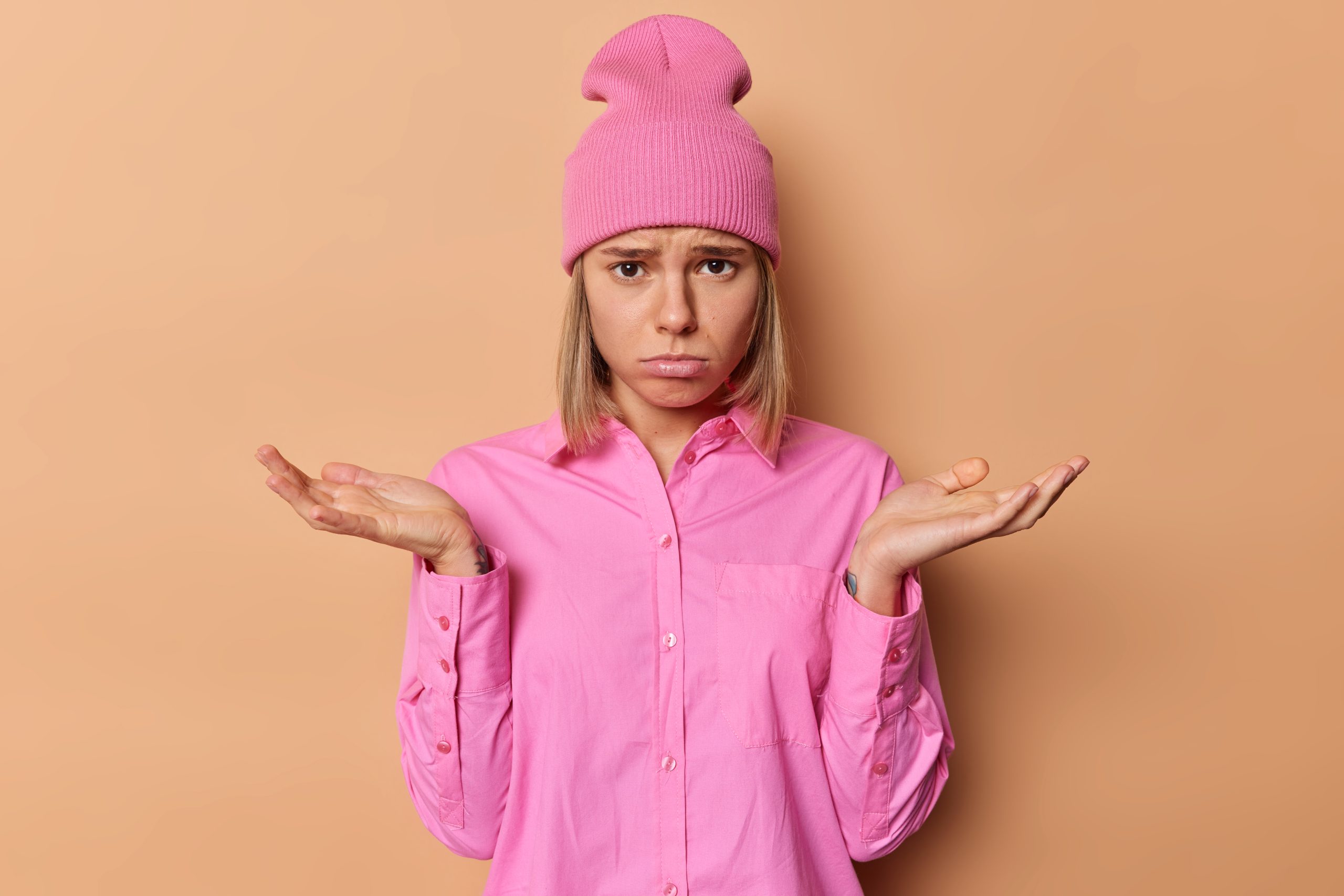 Displeased unhappy woman with fair hair looks sadly spreads palms has no answer stands clueless dressed in casual pink shirt and hat poses against brown background. Doubt and hesitation concept